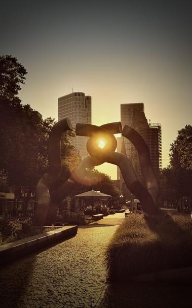 Das Bild zeigt eine moderne Skulptur in einem Stadtpark, wobei die Sonne durch eine Öffnung im Kunstwerk scheint. Im Hintergrund sind die umliegenden Gebäude zu sehen, die zu einer Sonnenuntergangsstimmung beitragen. Wege und Grünflächen unterstreichen die Parklandschaft.