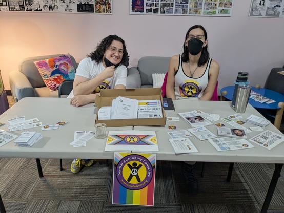 Perry and Allie of GAC behind a table full of stickers, pamphlets etc on genital autonomy.