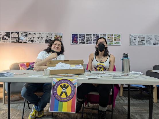 Perry and Allie of GAC behind a table full of stickers, pamphlets etc on genital autonomy.