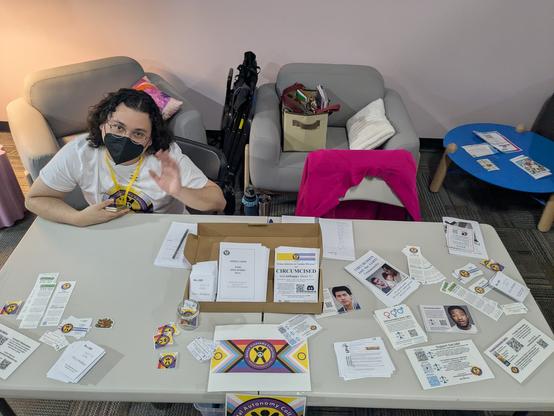 Perry of GAC behind a table full of stickers, pamphlets etc on genital autonomy.