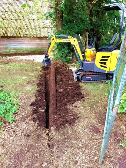 Mini excavator in black and yellow lifting a massive steel pipe out of the ground after uncovered it from shallow soil. The pipe continues towards the camera.
