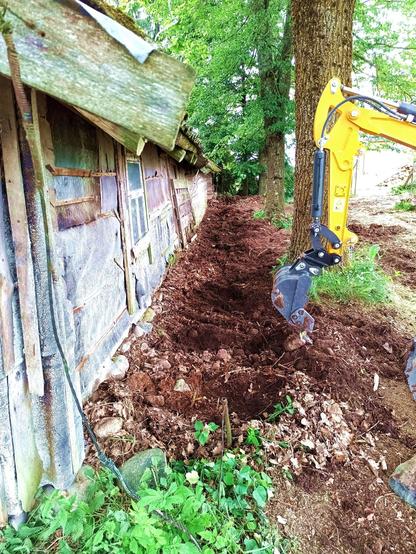 The side of an old log barn with a boarded up former door is being cleared from old junk, leaves, rocks, etc with a small excavator. The work happens between five large trees growing next to the barn. There's a window in the side of it.
