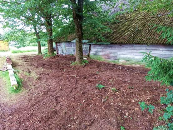 The area next to the barn afterwards. There's a white string spanning the length of it to help with the ditch gradient. A shallow ditch is made approximately where the rainwater from the roof drops off and there's a gradient away from the building into the ditch. The rest of the area is relatively level, apart from the far end which still needs filling in with more material. A massive concrete beam is laying on the left with a row of bricks stacked on top.