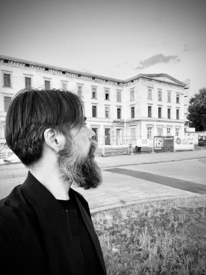 Ein Mann mit Bart steht im Profil und blickt auf ein großes, altes Gebäude. Das Bild ist in Schwarz-Weiß gehalten und zeigt einen teilweise bewölkten Himmel im Hintergrund. Die Umgebung scheint städtisch zu sein, mit Bauelementen und Gras im Vordergrund.