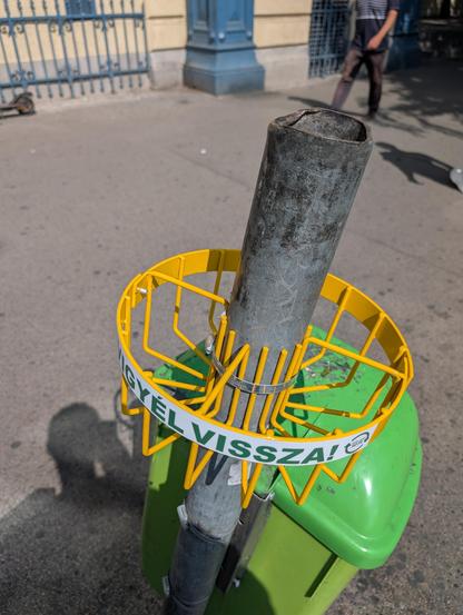 A round bottle holder, made up of thin bars of metal, is attached to a pole above a public trashcan right outside Nyugati railway station in Budapest. There's big label placed on the holder that says in Hungarian "take me back".