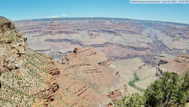 View from the South Rim of the Grand Canyon today. Blue sky, a few clouds on the horizon, and temperatures in the 70s. A trail that heads down towards the Colorado River appears as a faint line (center right). Trees dot the slopes in the foreground. A snaking line of vegetation can be seen in the lower right. Shade and natural water sources support plants and animals. Temperatures rise as one descends into the canyon, in summer and in winter months. From all angles, the colors of the rocks dominate the scene.