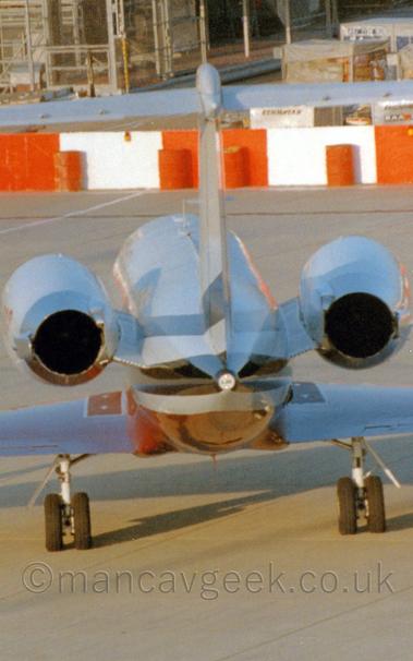 Rear shot of a twin engined BizJet parked on concrete apron facing away from the camera.
The plane is mostly white, with red and black stripes running along the body, under the engines mounted on the sides of the rear fuselage.
There are red and white barricades in the background, forming the perimeter of a building site.