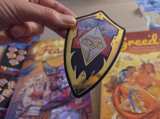 A sticker is held up close to the camera. It is shaped like a shield, with stripes of blue, pink and purple. Golden trim around it's edge and a golden heart with the infinity sign across in the center of the shield. The gold is reflecting the light and is very shiny. 
In the blurry background you can see the Feast and Greed books.