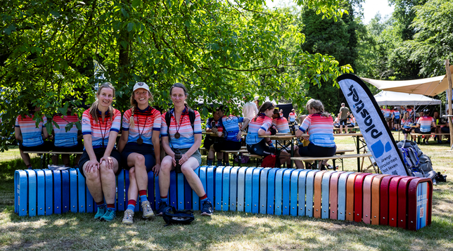 Three cyclists on a long bench with vertical elements in the colors of the climate stripes, presumably for the Netherlands.