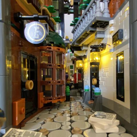 An image taking a close look at the Sherlock Holmes Booknook. The grey cobbles reflect the lamplight coming from the two lanterns on the right hand side white outside wall of 222B Baker Street. A pile of newspapers sits on the cobbles at the front right of the set. A bay window sticks out into the alley on the left of the view. A collection of books can be seen in the window display. A round sign with an ‘E’ hangs above the door to the Elementary Bookshop.