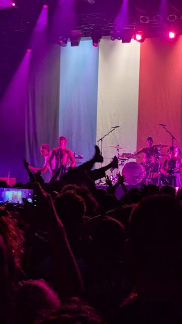 Eine Band auf der Bühne ist in violettes Licht getaucht. Im Vordergrund Publikum beim Crowdsurfen.