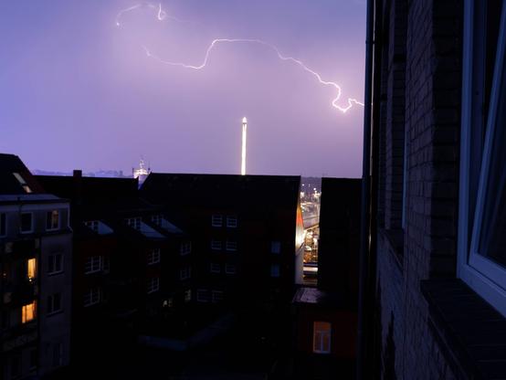 Eine Nachtaufnahme. Im Vordergrund sind dunkle Häusersilhouetten zu sehen, dahinter ein beleuchtetes Karussell, darüber ein Blitz.