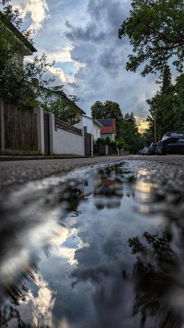 Das Bild zeigt eine ruhige Straße in einem Wohngebiet, auf der sich eine schmale Pfütze im Vordergrund befindet, in der sich Häuser, Bäume und ein Himmel mit dramatischen Wolken spiegeln. Auf der linken Straßenseite stehen gepflegte Häuser mit Vorgärten und Zäunen, während rechts einige Autos parallel zur Straße geparkt sind. Üppiges grünes Laub säumt beide Seiten.