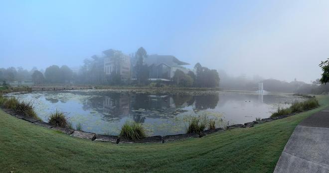 Panorama over the main UQ Lake. Thin fog softens the scene of a fountain with buildings and trees emerging from the fog in the distance.