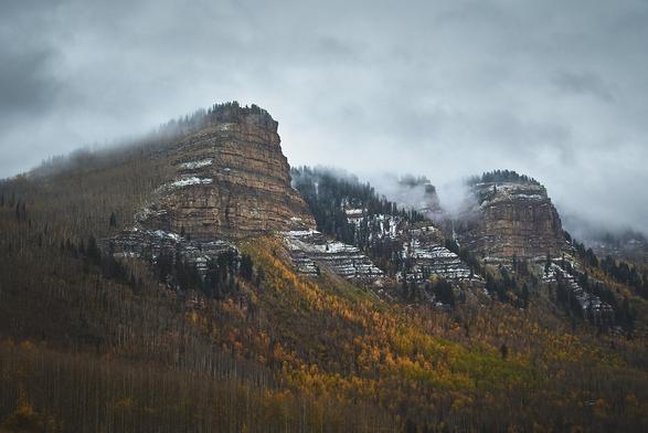 Misty mountain peaks with light snow, surrounded by autumn-colored forests under a cloudy sky.