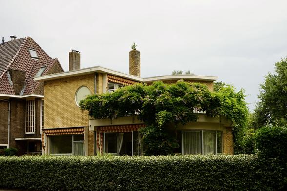 Vue de l'extérieur. Petite maison en brique jaune de style moderniste à moitié cachée par la végétation. Elle comporte une partie cubique à gauche et une partie arrondie avec des baies vitrées à droite.