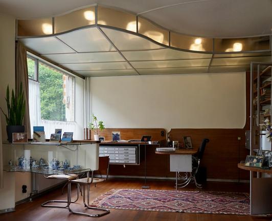 Vue du bureau depuis le salon. Le plafond translucide dessine des ondulations, des lumières à l'intérieur sont allumées. Au fond, on voit le bureau aux lignes très sobres, dont une partie est constituée de tiroirs fin renfermant des plans d'architecte. Une vitrine contenant une collection de porcelaine se trouve sur la gauche. Il y a aussi quelques chaises en acier tubulaire. Un tapis à motifs géométriques protège le parquet.