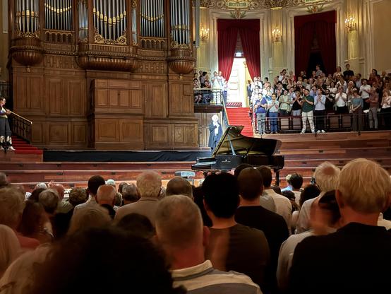 A grand concert hall filled with an audience applauding. A pianist stands next to a grand piano on stage, with an elaborate organ in the background. The audience is mostly seated, with a few people standing, and ornate decorations adorn the space.