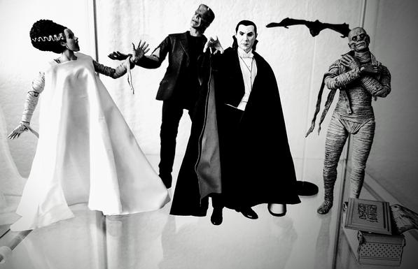 A black and white photo of a shelf display of NECA Ultimate Action Figures, including black & white versions of Dracula, Frankenstein's Monster, the Bride, and The Mummy.