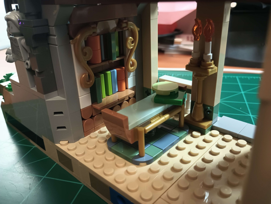 Closeup of Lego build, the inside of the bottom floor of a tower. A simple full-size bed with two pillows sits next to an ornate bookshelf with a dozen books, with spines of different greens, blues, and oranges. A tall candelabra with two candles sits next to the bed, next to a pillar.