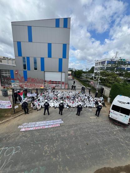 Une photo montrant plus d'une centaine de personnes assises habillées de blanc et masquées, bloquant ainsi le portail d'accès à un immeuble dans une zone industrielle. Elles sont entourées par une vingtaine de policiers debouts et casqués.
Au sol en premier plan, le slogan sur fond blanc: 
ELBIT SYSTEM SYENSQO
COMPLICE D'UN GENOCIDE
STOP ARMING ISRAEL