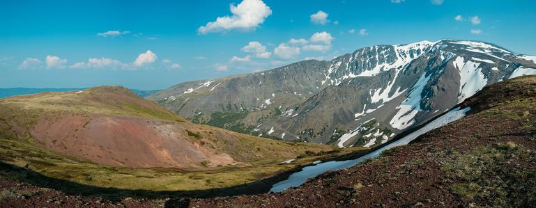 A stitched panorama from the top of an alpine meadow looking out toward more mountains, meadows, and the valley to the trail below. The mountains are snow capped and the sky is blue with a few clouds.