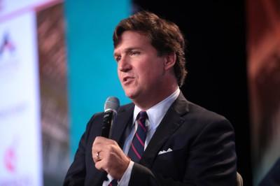 Tucker Carlson speaking into a microphone. Wearing a black jacket, white shirt and a tie