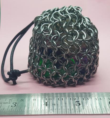 Steel Chainmail dice bag containing multicoloured dice against a pink backdrop.
A steel rule across the base