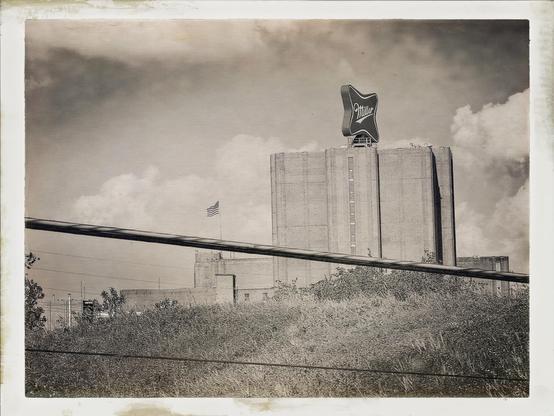 A vintage-style photograph of a large industrial building featuring a prominent sign that reads "Miller." An American flag is visible on the structure, set against a cloudy sky. In the foreground, tall grass and utility lines are present.