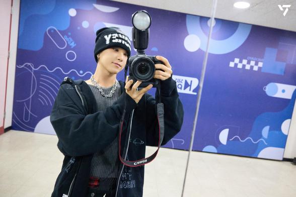 Hoshi takes a selfie in front of a mirror, wearing a black beanie, a dark hoodie, and a silver chain. He holds a camera with an attached flash, and colorful abstract art decorates the background wall.