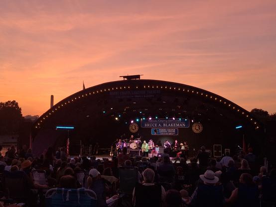 Picture of an amphitheater with a 7npiece band playing under an orange sunset on the hottest day of the year, so far.