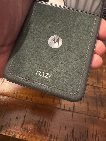 Human hand holding a cellphone so you can see the back of it. It's a dark green color and it looks to me made of a suede/leather material with the Motorola logo and "Razr" etched into it