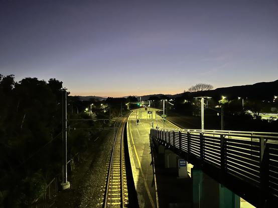 A train station at dawn, featuring empty tracks illuminated by lights. The platform is visible with a pedestrian bridge overhead, surrounded by trees and mountains in the background. The sky transitions from dark blue to orange at the horizon. A crescent moon is barely visible.