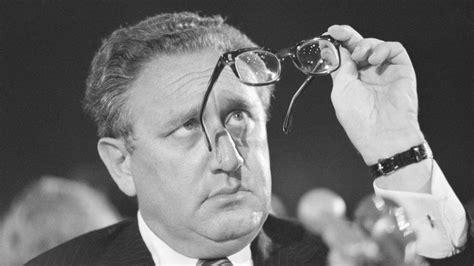 Blank and white photo of Henry Kissinger looking at his glasses.