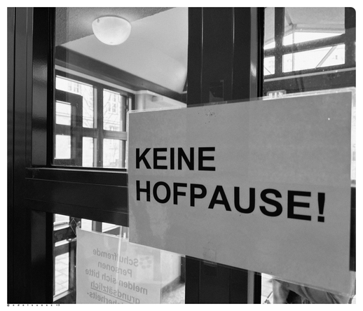Schwarzweißfotografie: eine alte Holztür mit großen Scheiben in einer Schule, darauf eine geklebter Ausdruck in dunklen Großbuchstaben: „KEINE HOFPAUSE!“ für die Schülerinnen, dahinter ein Foyer mit großer leuchtenden Deckenlampe