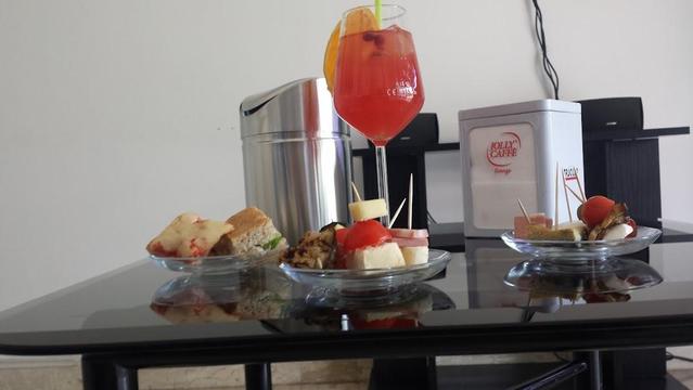 A glass of red cocktail sits on a glossy black table with plates of hors d'oeuvres, a silver shaker, and a JOLLY CAFFE container.