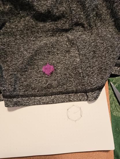 The bottom of a mottled black and gray skirt, with a small purple hexagon embroidered on it in satin stitch. The skirt is atop a piece of paper on which the same size hexagon is inscribed in a circle (which was used to construct the hexagon).