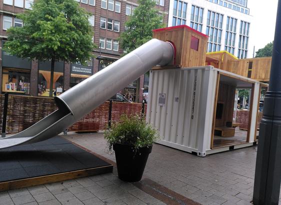 A small playground built in, on and around a freight container. The picture features prominently a covered slide and a small potted plant beside it. High city buildings in the background.