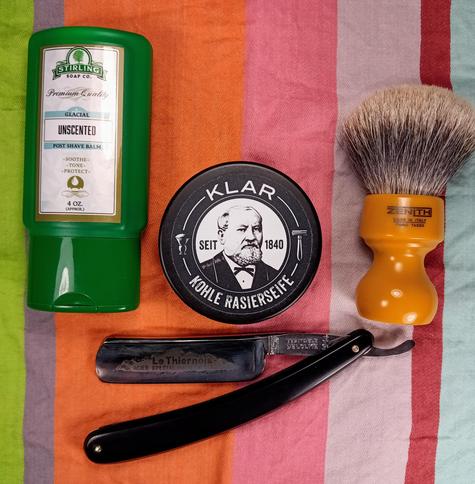 (Le Chat Mistral description)

The image showcases a collection of shaving items arranged on a striped background. On the left, there is a green container of Stirling Soap Co. unscented post-shave balm. In the centre, there is a round tin of Klar shaving soap, featuring an old-fashioned portrait and text in German. To the right of the tin, there is an orange-handled shaving brush with the brand name Zenith. Below the tin, there is a black straight razor with the brand name "Le Thiernois" engraved on it. The items are neatly laid out, suggesting a traditional wet shaving setup.