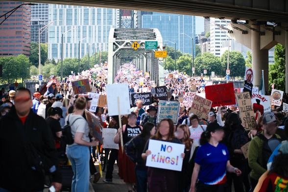 A large crowd of people marches across the Hawthorne Bridge in Portland, Oregon, under a clear sky. Many hold signs with various messages, some visible, like "NO KINGS!" in the foreground. The bridge stretches into the background, packed with more participants, and the office buildings lining Naito Parkway in downtown Portland are visible on either side of the bridge in the distance. Some trees line the riverbanks. The march appears to be busy and well-attended.