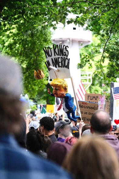 A large papier-mâché figure of Donald Trump in a blue suit and red tie hangs from a pole, grasping at a crown suspended on a wire just out of his reach. Above him, a sign reads "NO KINGS IN AMERICA". The figure is positioned above a crowd of protestors at the No Kings rally and march in Portland, OR, and in the background, the mast of the USS Oregon memorial is visible, along with green trees and more American flags.