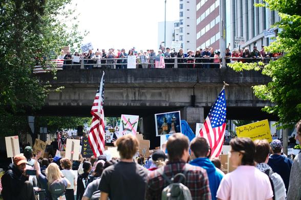 A large crowd of people gathers for the No Kings rally and march in Portland, OR, with many holding American flags and various signs. Above the underpass, another group of people stands on a pedestrian walkway or overpass, looking down at the marchers, some also holding signs or flags. Office buildings lining Naito Parkway are visible in the background, and green trees frame the sides of the image.