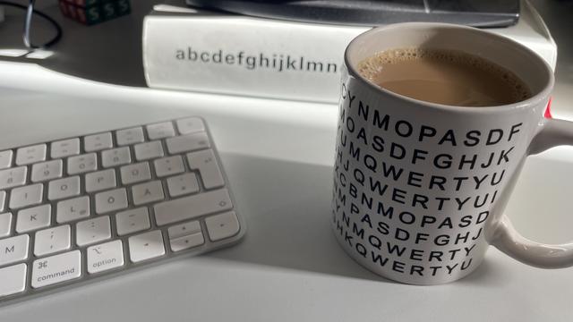 Das Bild zeigt eine weiße Schreibtischszene mit einer Tastatur im Vordergrund, einer Kaffeetasse mit schwarzem Buchstabenmuster im QWERTY-Stil und einem Buch im Hintergrund mit der Aufschrift „abcdefghijklmnopqrstuvwxyz“. Der Kaffee in der Tasse ist hellbraun, vermutlich mit Milch. Die Szene ist von Sonnenlicht durchflutet.