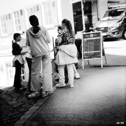 Schwarzweißfoto von 5 Mädchen, die in einer Fußgängerzone vor einem Geschäft im Kreis stehen und miteinander reden. Diana F+-Spielzeugkamera auf Ilford XP2 400-Film.