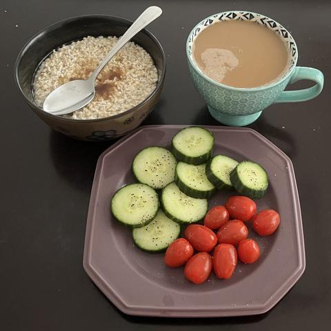 A plate of sliced cucumbers and cherry tomatoes seasoned with salt and pepper, a large bowl of oatmeal topped with brown sugar, and a cup of almond coffee.