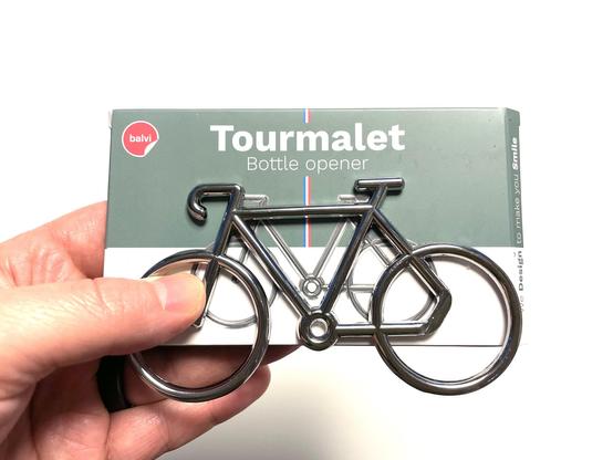 Kädessä polkupyörän muotoinen metallinen pullonavaaja, takana pahvilaatikko tekstillä ”Tourmalet bottle opener”.