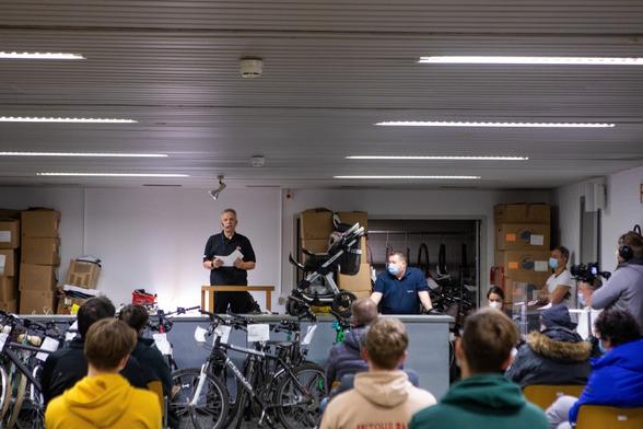 Ein Raum voll mit Fahrrädern, Kartons und zwei Personen im Vordergrund an einem Tisch stehend, die Auktion der Fundsachen leitend. Mehrere Menschen sitzen und schauen in deren Richtung. Ein Mann auf der rechten Seite filmt die Szenerie mit einer Kamera. Fahrradreifen und ein Kinderwagen sind im Hintergrund sichtbar.
© Giacomo Zucca, Bundesstadt Bonn