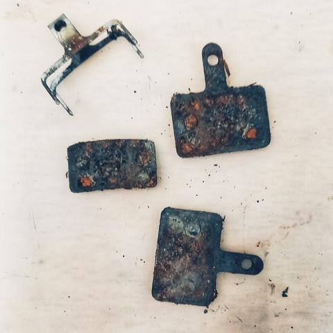 Une paire de plaquettes de frein de vélo, de forme rectangulaire, posées sur une surface claire. Elles sont rongées par la rouille et la crasse.
Quatre éléments sont visibles : en haut à gauche une sorte de pince (le ressort de rappel des plaquettes), juste en-dessous un petit rectangle noir et rouillé détaché des plaquettes, puis sur la droite l'une en dessous de l'autre les plaquettes elles-mêmes, rectangulaires avec une tige percée.