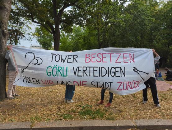 Ein Banner auf dem steht Tower besetzen, Görli verteidigen, holen wir uns die Stadt zurück. Außerdem ist ein Bolzenschneider abgebildet und ein Anker der symbolisch dafür stehen könnte, Zäune zu überwinden oder den Tower zu erklimmen. Dahinter ist ein Stück von der Versammlung vor dem Görlitzer Park zu sehen.