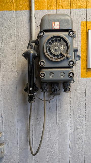 Foto eines sehr alten, wohl nicht mehr funktionsfähigen Wandtelefons.  Der Apparat ist grau, der Hörer auf der linken Seite schwarz, die Wand weiß gestrichen und oben rechts sieht man gelbe Farbe.